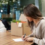 Donna che scrive lettera di dimissioni al lavoro con scatola di trasloco in ufficio